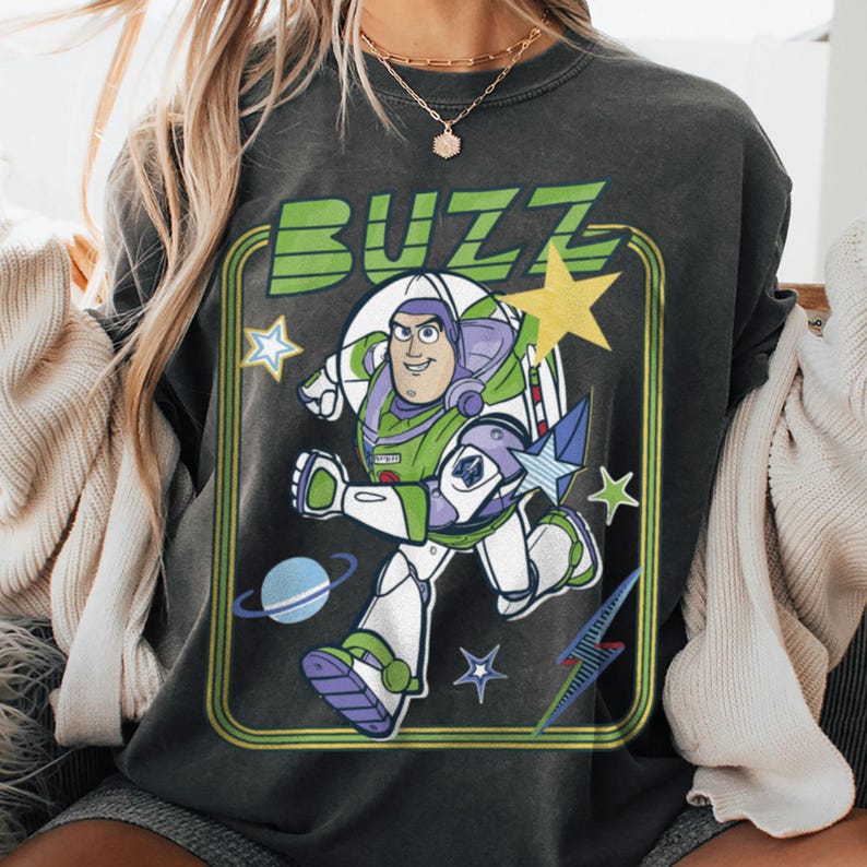 May include: A charcoal grey t-shirt featuring a graphic print of the character Buzz Lightyear in a running pose. The design includes the word BUZZ in green letters, a yellow star, a blue planet with rings, and smaller star shapes inside a green frame.