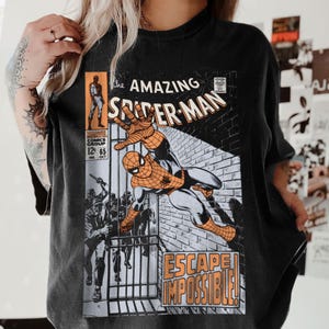 May include: Black t-shirt featuring a vintage comic book cover of Spider-Man. The graphic includes the title "The Amazing Spider-Man" and the phrase "Escape Impossible!" in orange and white. The comic book cover art depicts Spider-Man in action.