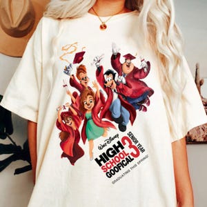 May include: Cream-colored t-shirt featuring a colorful graphic of Disney characters in graduation attire. The design includes the text "High School Goofical Senior Year 33 Graduating This Spring!"