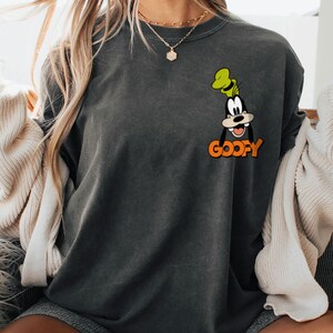 Puede incluir: Sudadera gris oscuro con una imagen de dibujos animados de Goofy con un sombrero verde y la palabra "Goofy" en naranja. La sudadera es de cuello redondo y tiene un ajuste relajado. La imagen est&aacute; en la parte delantera de la camisa.