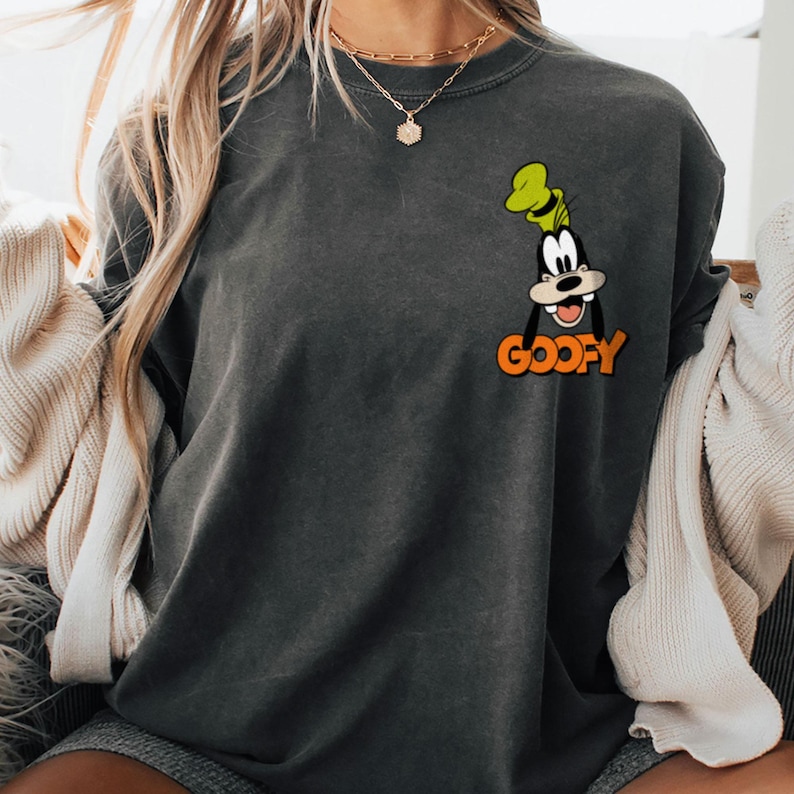 Puede incluir: Sudadera gris oscuro con una imagen de dibujos animados de Goofy con un sombrero verde y la palabra "Goofy" en naranja. La sudadera es de cuello redondo y tiene un ajuste relajado. La imagen est&aacute; en la parte delantera de la camisa.