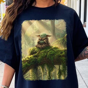 May include: Navy blue t-shirt with a detailed illustration of a green alien-like character, seated on a moss-covered log in a forest. The character is wearing a brown robe and has its hands clasped. The background shows a lush forest with trees.