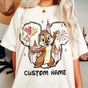 May include: Cream-colored t-shirt featuring a cartoon illustration of Chip from Chip 'n Dale Rescue Rangers. The design includes a castle, balloons, fireworks, and the text "CUSTOM NAME" in black. The shirt has short sleeves.
