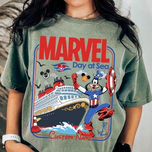 May include: Sage green t-shirt featuring a Marvel Day at Sea design. The graphic includes a cruise ship, Captain America Goofy, and the text "MARVEL Day at Sea" in red and blue. The shirt has a vintage, distressed look.