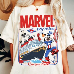 May include: Cream-colored t-shirt featuring a Marvel Day at Sea design. The graphic includes a cruise ship, Captain Goofy, and the text "MARVEL Day at Sea" in red and blue. The design also includes Mickey Mouse icons and the text "Custom-Name".