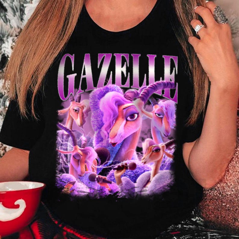 May include: Black t-shirt featuring a purple graphic of cartoon gazelles with microphones. The word "GAZELLE" is printed in a purple gradient font above the gazelles. The gazelles have purple hair and are singing.