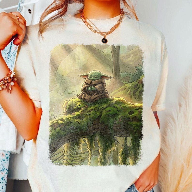 May include: A cream-coloured t-shirt with a detailed illustration of a green alien character from a popular science fiction series. The character is seated on a moss-covered log in a forest setting. The artwork has a soft, painterly style.