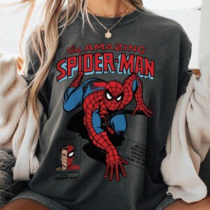 以下が含まれることがあります： スパイダーマンのコミックイラストが描かれた濃いグレーのスウェットシャツ。「the AMAZING SPIDER-MAN」の文字が赤と青で描かれ、ピーター・パーカーの小さな画像も含まれています。スーパーヒーローは、彼のクラシックな赤と青のスーツで描かれています。