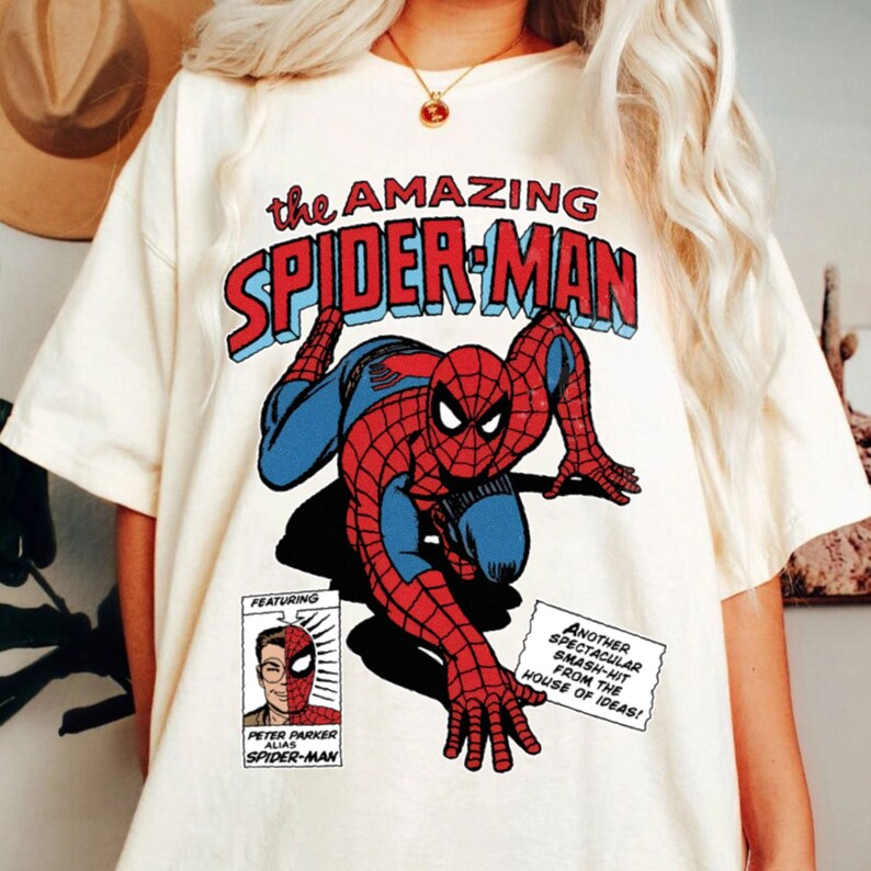 Może przedstawiać: Kremowa koszulka z komiksową grafiką Spider-Mana w kolorze czerwonym i niebieskim. Tekst "the AMAZING SPIDER-MAN" znajduje się nad obrazem. Dodatkowy tekst zawiera "FEATURING" i "PETER PARKER ALIAS SPIDER-MAN".