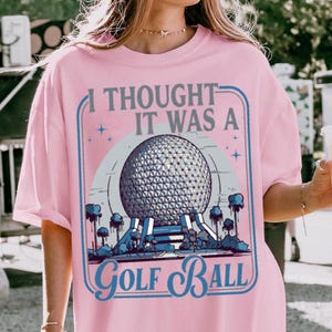 May include: Pink t-shirt with a graphic that reads "I Thought It Was A Golf Ball." The graphic features a geometric sphere design with a blue and white color scheme, and a blue border.