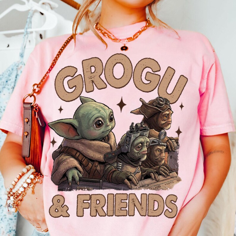 K&ouml;nnte beinhalten: Hellrosa T-Shirt mit dem Text "GROGU & FRIENDS" in braunen Buchstaben. Die Grafik zeigt eine gr&uuml;ne Grogu-Figur und drei weitere Alien-Figuren. Das Design ist vor einem rosa Hintergrund.