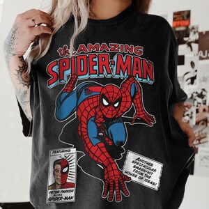 Może przedstawiać: Czarny t-shirt z grafiką w stylu vintage komiksu Spider-Mana w kolorze czerwonym i niebieskim. Tekst "the AMAZING SPIDER-MAN" znajduje się na g&oacute;rze. Dodatkowy tekst to "FEATURING PETER PARKER ALIAS SPIDER-MAN" i "ANOTHER SPECTACULAR SMASH-HIT FROM THE HOUSE OF IDEAS!"