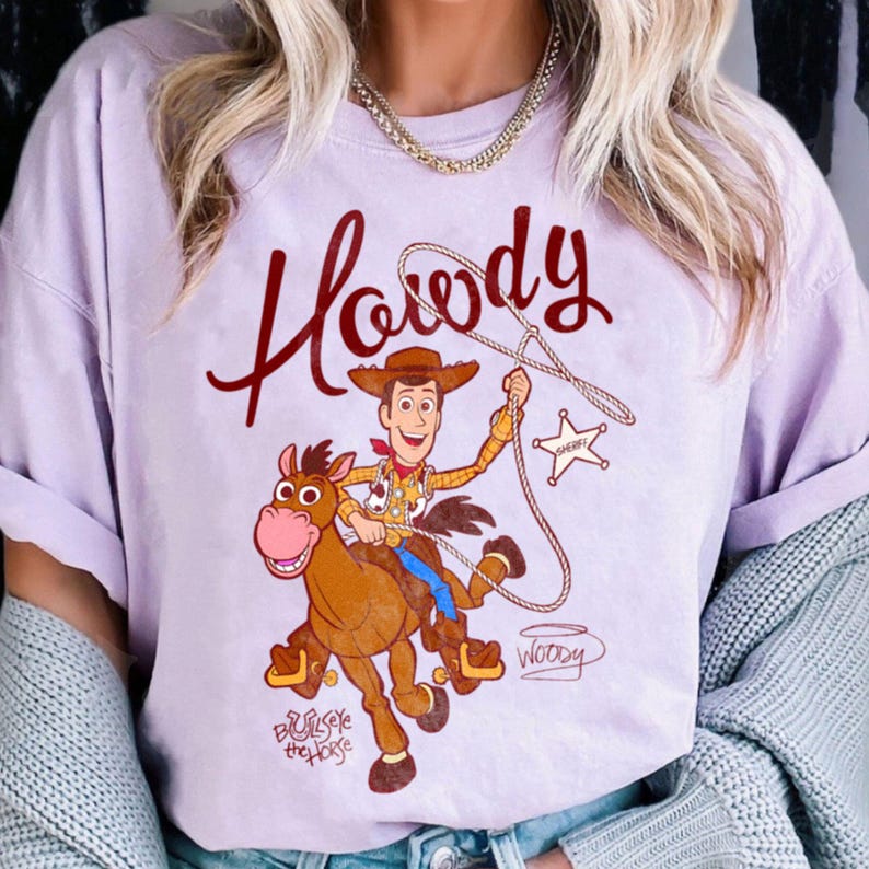 May include: A light purple t-shirt featuring a cartoon image of Woody from Toy Story riding Bullseye the horse. The word "Howdy" is written in a cursive font above the image. The shirt also has the words "Sheriff" and "Woody" on it.