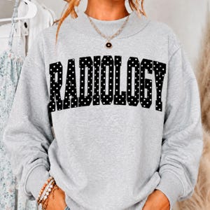 May include: A light gray crewneck sweatshirt featuring the word RADIOLOGY printed in a bold, black, block-style font with white polka dots throughout the letters. The garment has long sleeves and a ribbed neckline, cuffs, and hem.