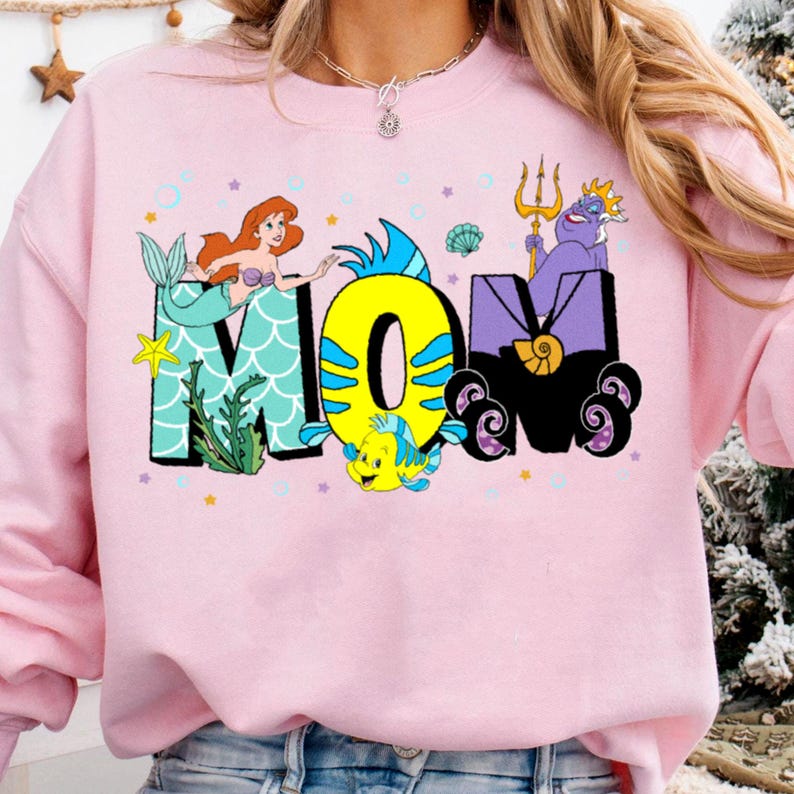 May include: Pink sweatshirt with a graphic design of the word "MOM" in a whimsical font. The letters are decorated with images of Ariel from The Little Mermaid, Flounder, and Ursula.