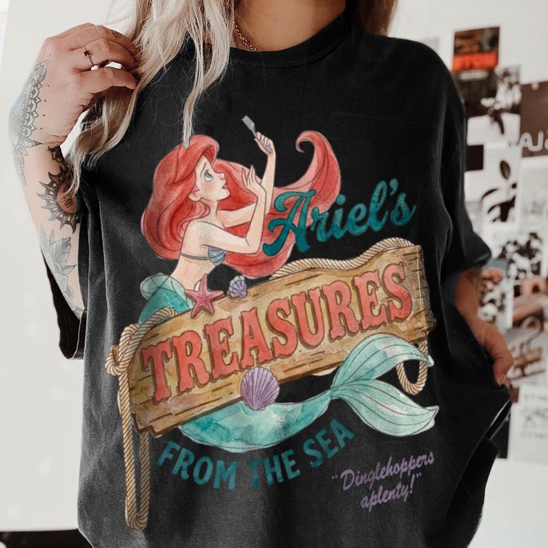 May include: Black t-shirt featuring a watercolor illustration of Ariel from The Little Mermaid. The design includes the text "Ariel's Treasures From The Sea" on a wooden sign, with a mermaid tail and the phrase "Dinglehoppers aplenty!"