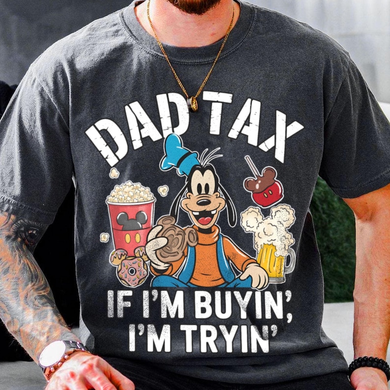 Op de afbeelding: Donkergrijs t-shirt met de tekst "DAD TAX" en "IF I'M BUYIN', I'M TRYIN'". Het shirt heeft een cartoonfiguur die snacks en een biertje vasthoudt. De figuur draagt een oranje shirt en een blauwe overall.