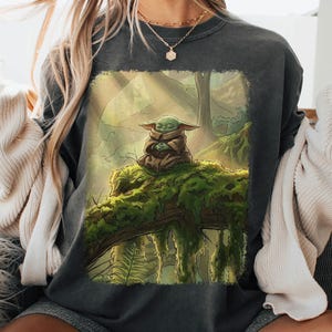 May include: Charcoal grey sweatshirt with a forest scene print. The design features a green alien character in a meditative pose on a mossy log. The artwork is framed by a soft green border, creating a whimsical design. The sweatshirt is a comfortable, casual garment.