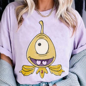 May include: A light purple t-shirt featuring a cartoon alien with one eye, a yellow body, and a smiling face. The alien has a long, curved antenna and two heart-shaped teeth. The shirt has rolled-up sleeves.