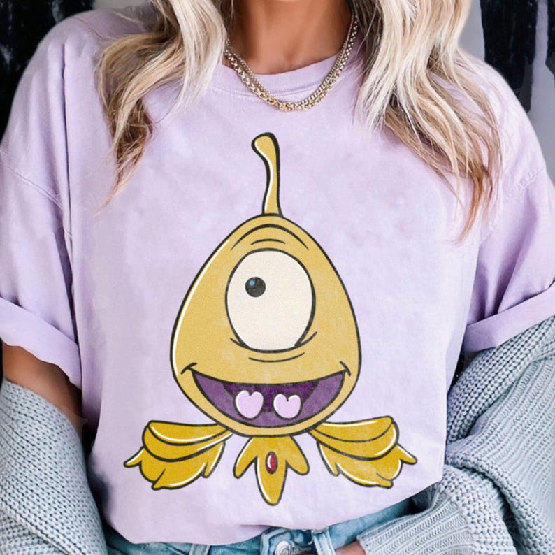 May include: A light purple t-shirt featuring a cartoon alien with one eye, a yellow body, and a smiling face. The alien has a long, curved antenna and two heart-shaped teeth. The shirt has rolled-up sleeves.