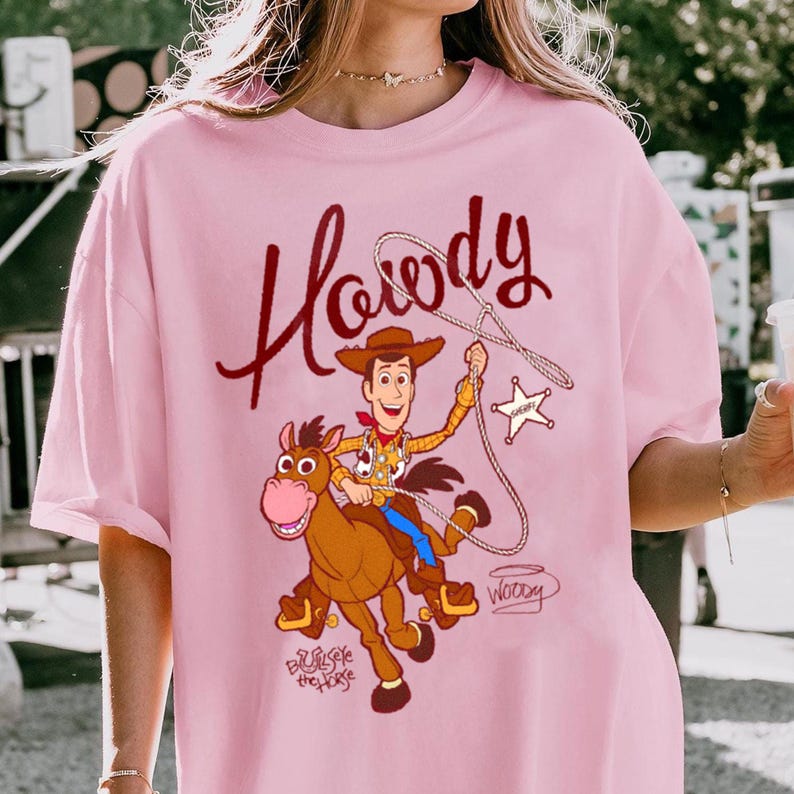 May include: Pink t-shirt featuring a cartoon illustration of Woody from Toy Story riding a horse. The shirt has the word "Howdy" in a script font above the illustration. The horse is brown, and Woody is wearing a cowboy hat and vest.