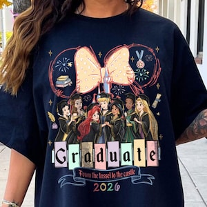 Puede incluir: Camiseta azul marino con un dise&ntilde;o de princesas de Disney vestidas con atuendos de graduaci&oacute;n. La palabra "Graduate" se muestra en letras de bloque coloridas, con el texto "From the tassel to the castle 2026" debajo. Un gran lazo y fuegos artificiales completan el dise&ntilde;o.