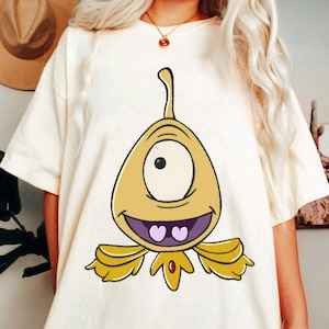 May include: Cream-colored t-shirt featuring a cartoon alien with one eye, a wide smile, and yellow and purple accents. The alien has a teardrop-shaped body and a curved antenna. The shirt is a casual, oversized fit.