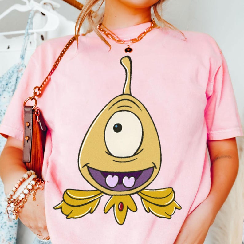 May include: Pink t-shirt featuring a cartoon character with a single eye, yellow body, and purple mouth. The character has a smiling expression and is centered on the shirt. The shirt is a solid pink color.