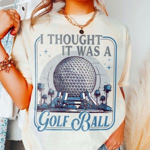 May include: Cream-colored t-shirt with a graphic design featuring a geometric sphere, stylized trees, and the text "I Thought It Was A Golf Ball" in blue. The design is framed by a blue border with stars.