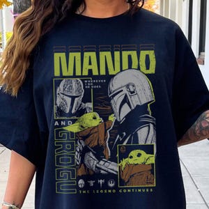 May include: Black t-shirt with a yellow graphic design featuring the words "MANDO" and "GROGU". The design includes images of a Mandalorian helmet and Grogu, with the phrase "Wherever I go, he goes." The bottom reads "The Legend Continues."