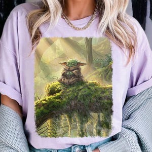 May include: A lilac t-shirt with a detailed illustration of a small, green creature from a popular science fiction series. The creature is seated on a moss-covered log in a forest setting, with a serene expression.