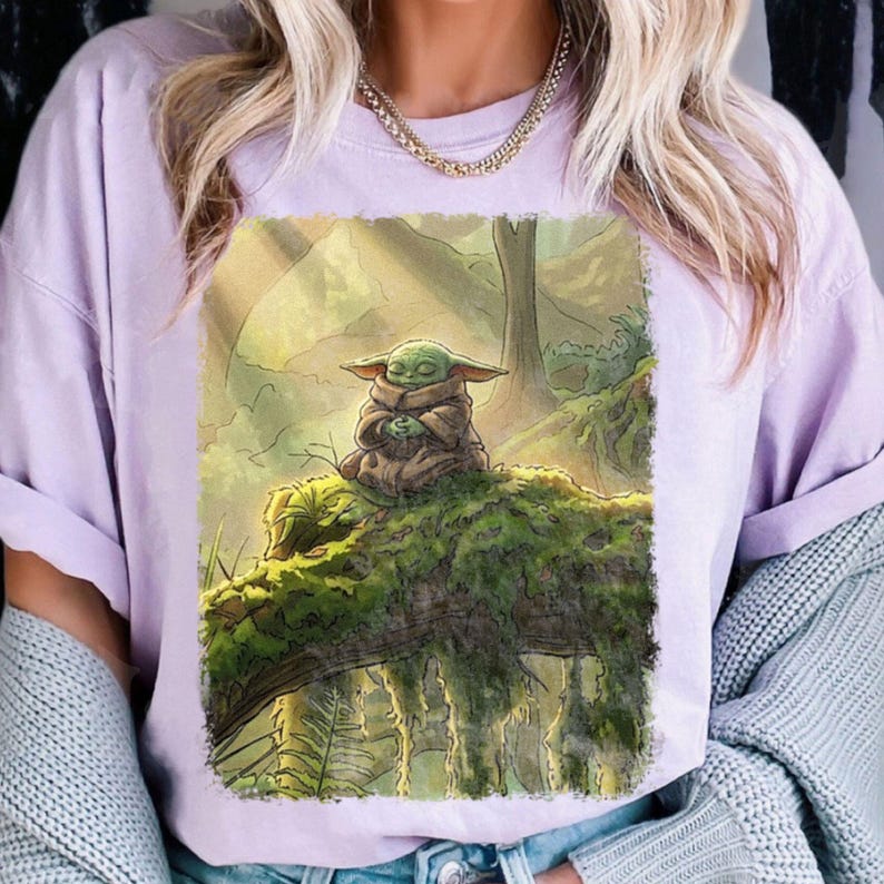 May include: A lilac t-shirt with a detailed illustration of a small, green creature from a popular science fiction series. The creature is seated on a moss-covered log in a forest setting, with a serene expression.