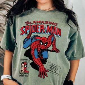 以下が含まれることがあります： セージグリーンのTシャツ。ヴィンテージコミックデザインのスパイダーマンが特徴です。「the AMAZING SPIDER-MAN」の文字が赤と青で描かれ、ピーター・パーカーのコミックストリップも含まれています。クラシックなスーパーヒーローTシャツです。
