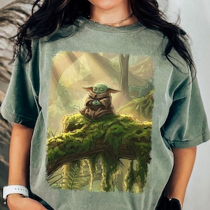May include: Sage green t-shirt with a graphic of a green alien-like character meditating on a moss-covered log in a forest. The artwork has a soft, painterly style with muted colours.
