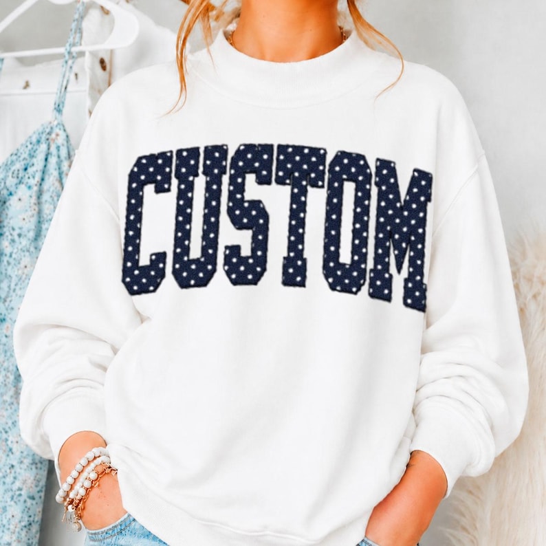 May include: A white long-sleeve sweatshirt featuring the word CUSTOM in large, dark blue block letters with a white polka dot pattern. The sweatshirt has a mock neck collar and long sleeves, providing a clean and simple look for casual wear.
