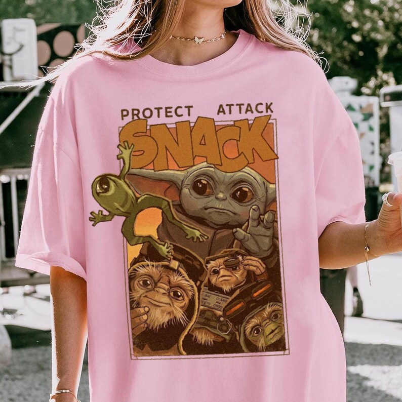 May include: Pink t-shirt featuring a graphic of a green alien, a character resembling Baby Yoda, and other creatures. The text "PROTECT ATTACK SNACK" is printed above the characters in a brown font. The shirt is a light pink color.