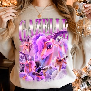 May include: Beige sweatshirt featuring a purple graphic of the word "Gazelle" above a cartoon image of gazelles with purple hair. The gazelles are holding microphones and are surrounded by purple lightning bolts.
