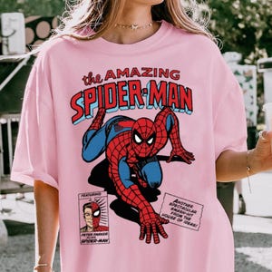 以下が含まれることがあります： スパイダーマンのコミックブックグラフィックが特徴のピンクのTシャツ。デザインには、赤と青の「the AMAZING SPIDER-MAN」の文字と、コミックブックパネルが含まれています。カジュアルウェアアイテムです。
