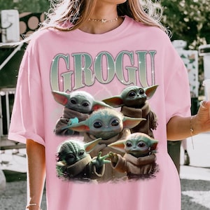 May include: Pink t-shirt with a graphic of Grogu, and the word "GROGU" above the image. The image shows five Grogu characters in different poses. The t-shirt is made of a soft material.