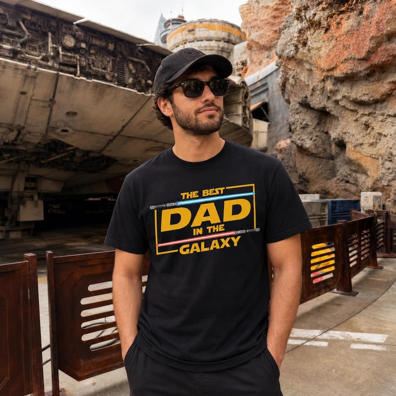 May include: Black t-shirt with the text "THE BEST DAD IN THE GALAXY" in a yellow, blue, and pink design. The shirt is worn by a person wearing a black cap and sunglasses.
