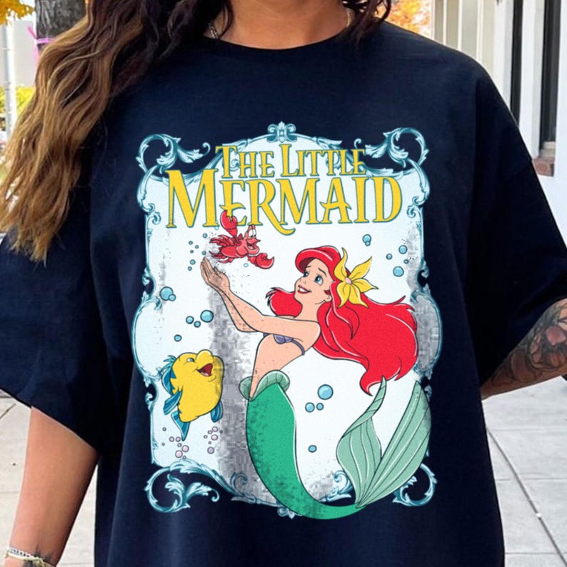 Puede incluir: Camiseta negra con un gr&aacute;fico colorido de "La Sirenita". El dise&ntilde;o incluye a Ariel con cabello rojo, una cola verde y una flor amarilla, interactuando con un cangrejo rojo y un pez amarillo. Las palabras "THE LITTLE MERMAID" se muestran arriba.