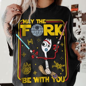May include: Black t-shirt with a humorous graphic featuring a fork character with lightsabers and a pacifier. The design includes the text "MAY THE FORK BE WITH YOU" in a yellow and red frame, with Star Wars-themed elements.