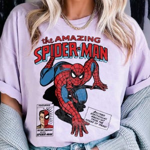 以下が含まれることがあります： スパイダーマンのコミック風イラストが描かれた、薄紫色のTシャツ。「the AMAZING SPIDER-MAN」の文字が赤と青で描かれ、ヒーローの別人格であるピーター・パーカーの小さな画像も含まれています。