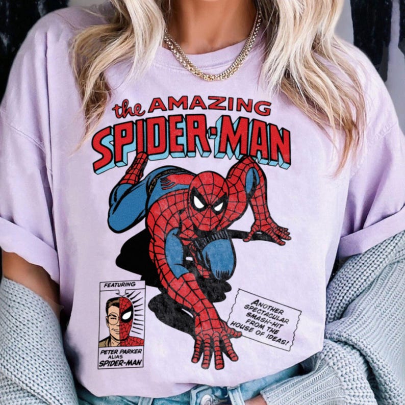 以下が含まれることがあります： スパイダーマンのコミック風イラストが描かれた、薄紫色のTシャツ。「the AMAZING SPIDER-MAN」の文字が赤と青で描かれ、ヒーローの別人格であるピーター・パーカーの小さな画像も含まれています。