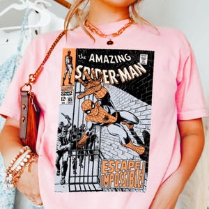 May include: A light pink t-shirt featuring a comic book cover of "The Amazing Spider-Man." The graphic shows Spider-Man in action with the text "ESCAPE IMPOSSIBLE!" and "MARVEL COMICS GROUP 12 65."