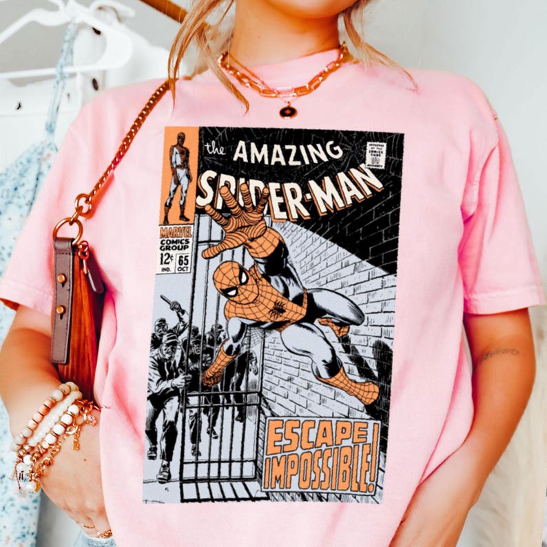 May include: A light pink t-shirt featuring a comic book cover of "The Amazing Spider-Man." The graphic shows Spider-Man in action with the text "ESCAPE IMPOSSIBLE!" and "MARVEL COMICS GROUP 12 65."