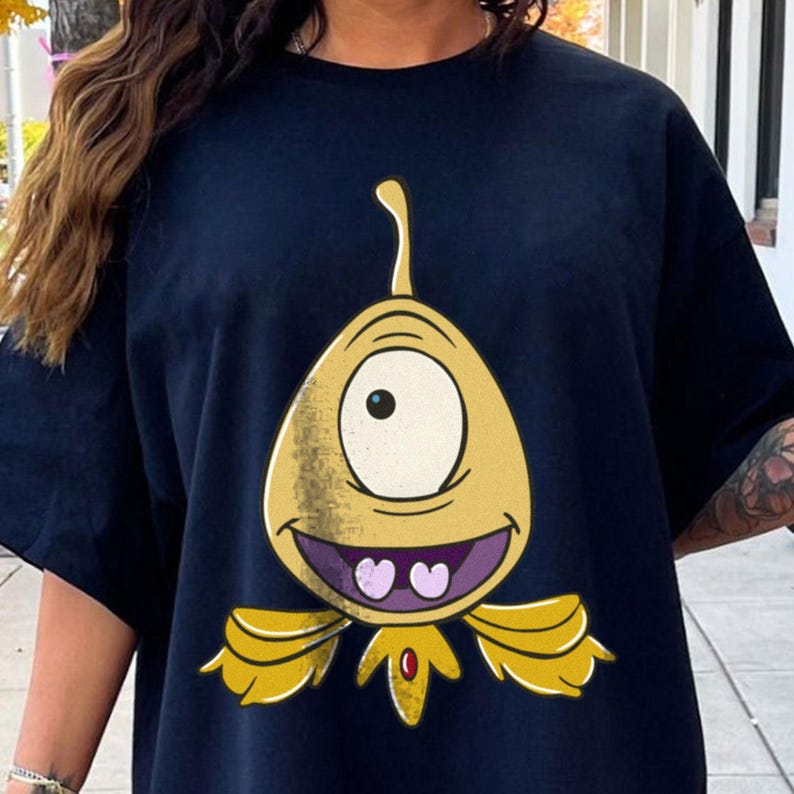 May include: Navy blue t-shirt featuring a cartoon alien with a large eye, yellow body, and purple mouth. The alien has two heart-shaped teeth and yellow wing-like appendages. The design is centered on the shirt.