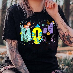 May include: Black t-shirt with a graphic of characters from the Disney movie The Little Mermaid. The graphic includes Ariel, Ursula, and Flounder. The text "MOM" is spelled out using the characters.