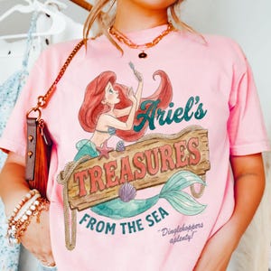 May include: Pink t-shirt featuring a graphic of Ariel from The Little Mermaid. The design includes Ariel, a wooden sign that reads "Ariel's Treasures From The Sea", and the phrase "Dinglehoppers aplenty!"