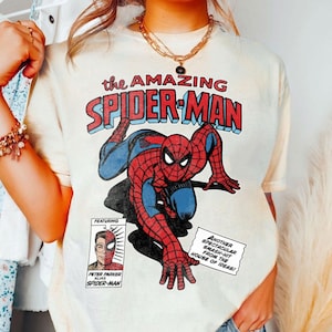 Może przedstawiać: Kremowa koszulka z komiksową ilustracją Spider-Mana. Projekt zawiera tekst "the AMAZING SPIDER-MAN" w kolorze czerwonym i niebieskim, a także dymki i portret Petera Parkera.